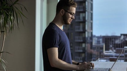 Man standing while using a laptop computer at his waist height like a goober.