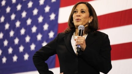 Kamala Harris holds a microphone and smiles in front of an American flag.