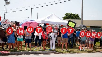 Protesters for the release of migrant children stand in front of a detention center with signs spelling out 'shut it down.'