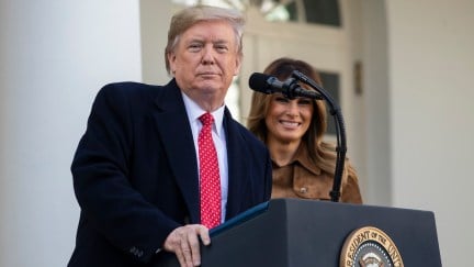 Donald Trump stands at a podium, in front of Melania, looking smugly at the camera.