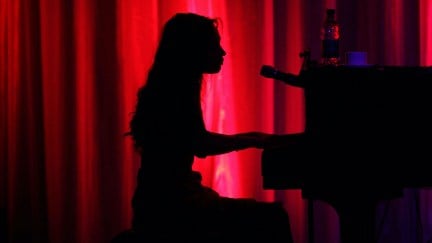 Fiona Apple plays piano in silhouette against a red curtain.