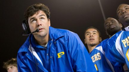 A white man in a coach uniform stands with his team looking off into the distance impatiently.