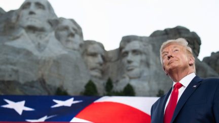 trumpstands in front of an image of mt. rushmore, smiling vacuously