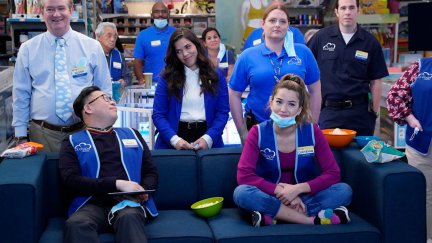 Characters from Superstore sit on and around a sofa, some in masks.