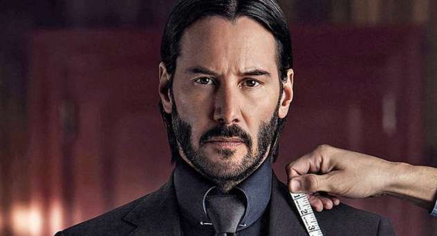 Keanu Reeves as John Wick looks into the camera as a tailor's hand holds a measuring tape to his collar