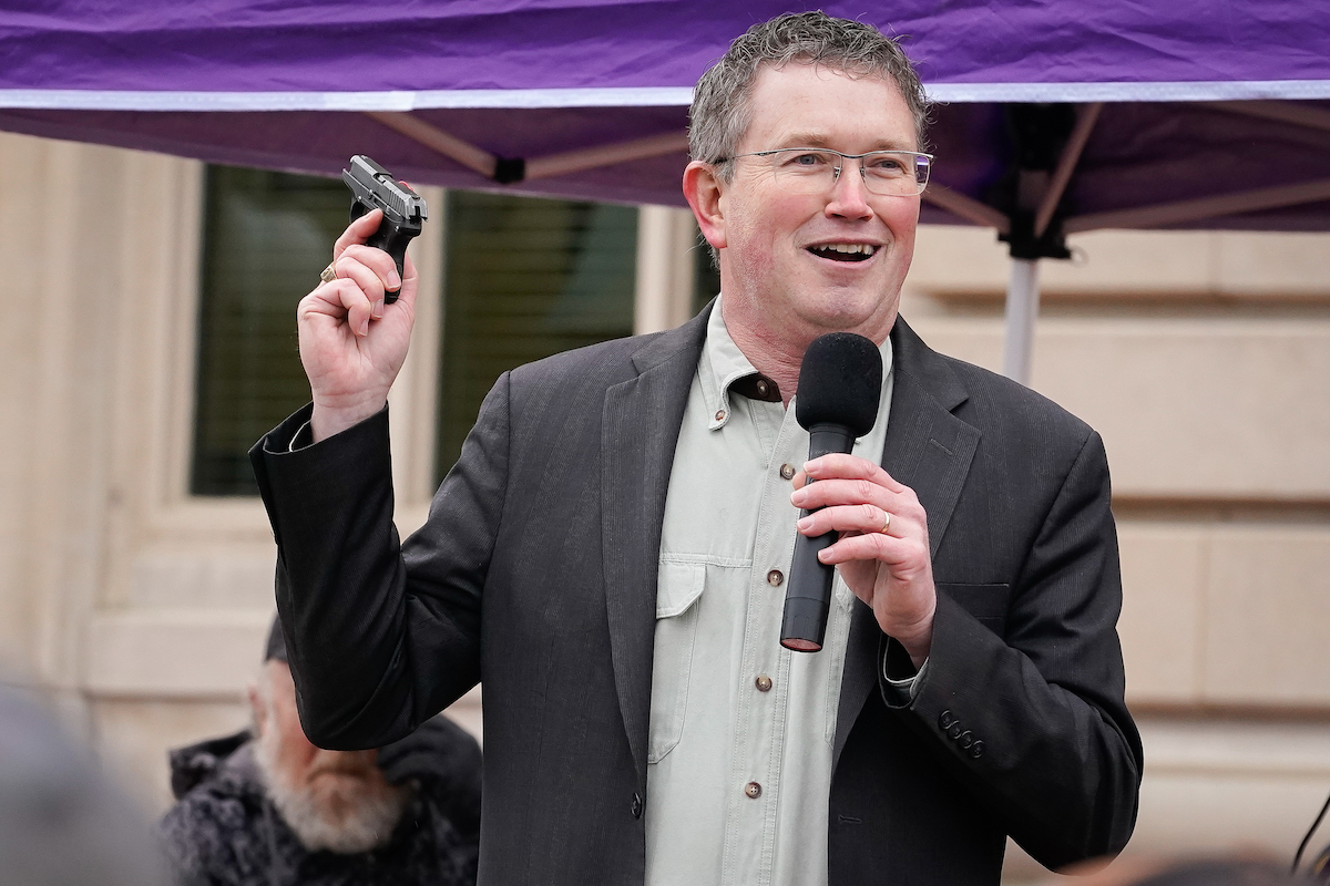 Thomas Massie holds up a small gun and smiles while holding a microphone during a rally.