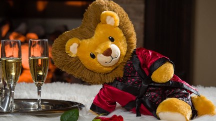 a stuffed lion in satin pajamas and robe is lounging suggestively next to glasses of champagne and a rose