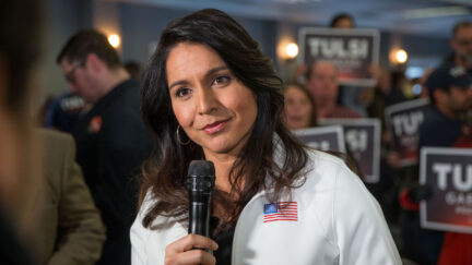 Tulsi Gabbard tilts her head and smirks, holding a microphone and speaking with a reporter