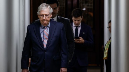 Mitch McConnell bites his lip and looks down as he walks with aides behind him.