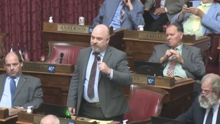 A middle aged white man stands and speaks, surrounded by other white men, in the West Virginia statehouse.