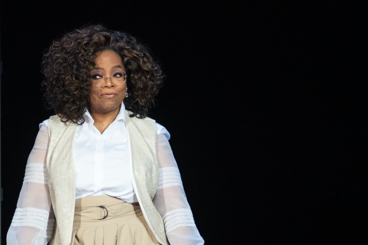 Oprah stands in front of a black background at a live event and smirks.