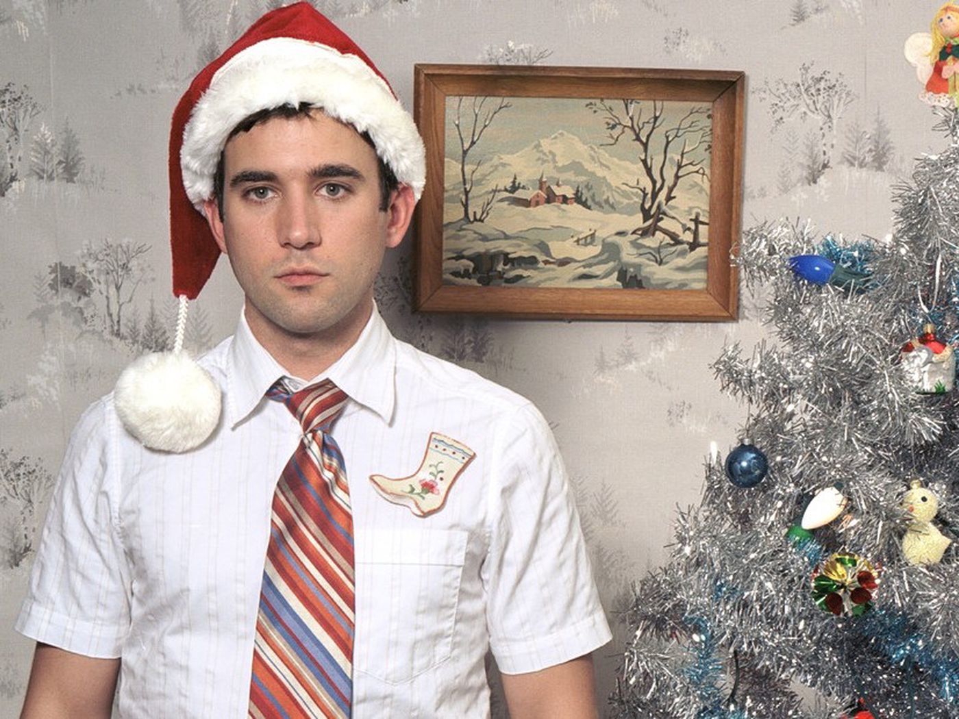 Sufjan Stevens wears a Santa hat and stands near a decorated Christmas tree