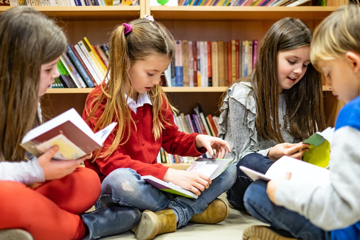 A group of young children sit on the floor of a library reading books.