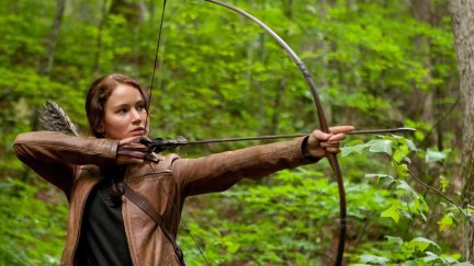 A brown haired teen girl with her hair in a braid pulls back her bow and arrow in the forest.