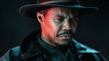 Publicity image of Gaius Charles as Perlie Armstrong on AMC's 'The Walking Dead: Dead City.' We see him from the shoulders up. He is a Black man wearing a wide-brimmed fedora/cowboy hat, a black leather jacket, and a denim button-down shirt. He has a mustache and the beginnings of a beard. He is scowling sternly and looking off to the side.