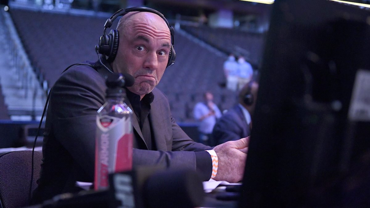 Joe Rogan mugs at the camera while sitting at a computer inside an arena, wearing headphones.