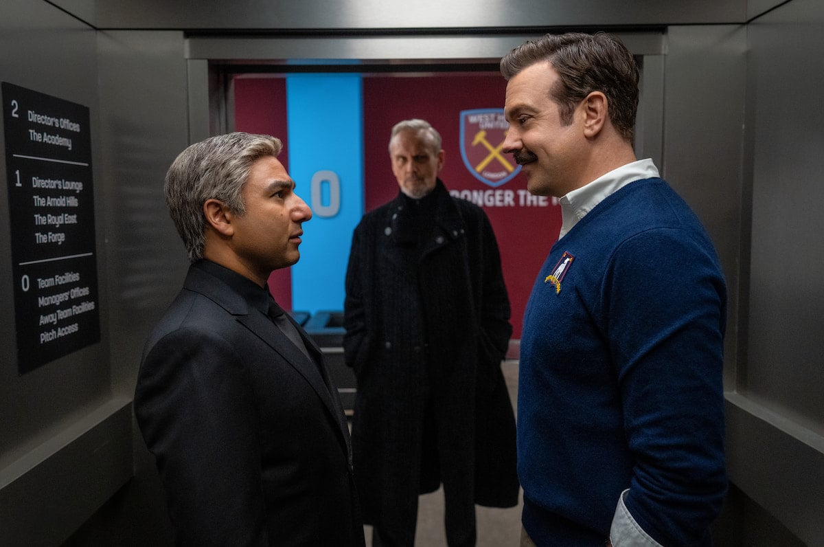 Nate and Ted stare each other down in a stadium hallway with Rupert looking on in the background.