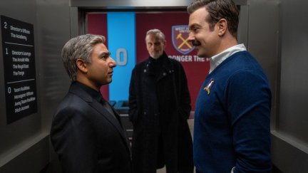 Nate and Ted stare each other down in a stadium hallway with Rupert looking on in the background.