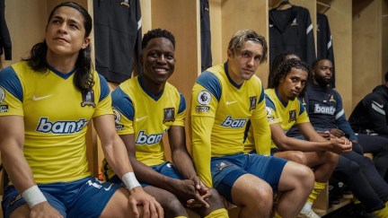 Cristo Fernández as Dani Rojas, Toheeb Jimoh as Sam Obisanya, Phil Dunster as Jamie Tarrt and Moe Hashim as Moe Bumbercatch sitting on a bench in a lockeroom, wearing their yellow and blue away game kits