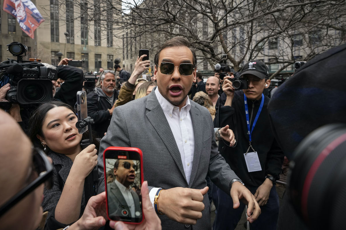 George Santos gapes open-mouthed, wearing sunglasses, surrounded by a crowd of reporters.