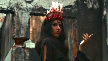 A woman with a black moon on her forehead and a headdress made of candles poses against a crumbling concrete structure.