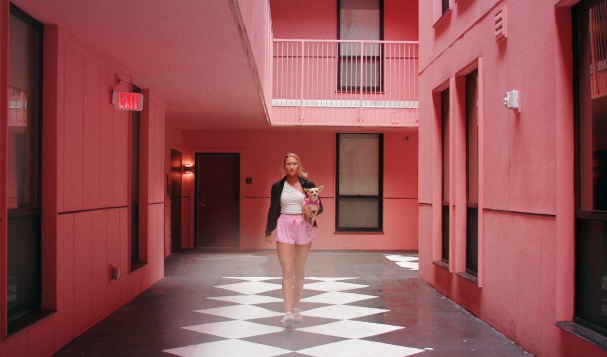 A young blonde woman walks down a pink-walled corridor carrying a small dog in 'Bama Rush'