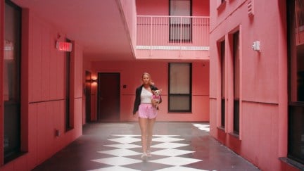 A young blonde woman walks down a pink-walled corridor carrying a small dog in 'Bama Rush'