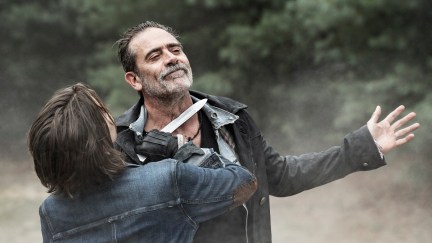 Lauren Cohan as Maggie Rhee and Jeffrey Dean Morgan as Negan in a scene from AMC's 'The Walking Dead: Dead City.' Maggie is a white woman with chin-length brown hair holding a knife to Negan's throat in her black fingerless gloves, wearing a denim jacket. Negan is a white man with dark hair and a salt-and-pepper 5 o'clock shadow wearing several layers of shirts under a black jacket. He's holding his arms out with smirk on his face accepting that a knife to his throat is how it's gonna be now. They're standing outside with grass and trees in the background.