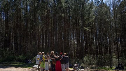 A group of about a dozen people embrace, with the tall trees of a forest behind them.