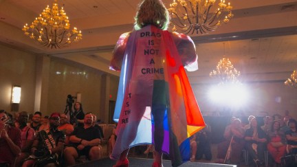 A drag performer is seen on a catwalk from behind, wearing a rainbow cape that reads 