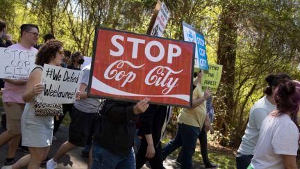 Protesters carry signs through a forest, including one that says 