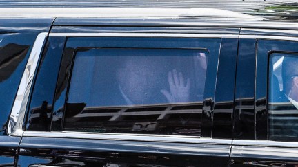 Donald Trump sits in the backseat of a car, waving through a tinted window.