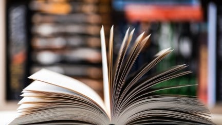 An open book on a table with book shelves in the background.