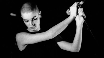 A black and white photo of a young Sinead O'Connor holding a microphone mournfully.