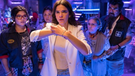 Milagro (Belissa Escobedo), Rocio (Elpidia Carrillo), Jenny (Bruna Marquezine), Nana (Adriana Barraza), and Uncle Rudy (George Lopez) in a scene from 'Blue Beetle.' Milagro, Rocio, Nana, and Rudy stand in a group behind Jenny, who is touching a device on her wrist.