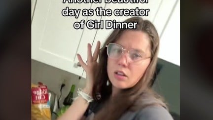 Screencap of a TikTok featuring @liviemaher. She is a young, white woman with long, brown hair wearing thin-rimmed glasses and a grey t-shirt. She's holding a hand to her ear as she holds her phone up in her other hand to shoot a video as she stands in her kitchen. The text above her reads 