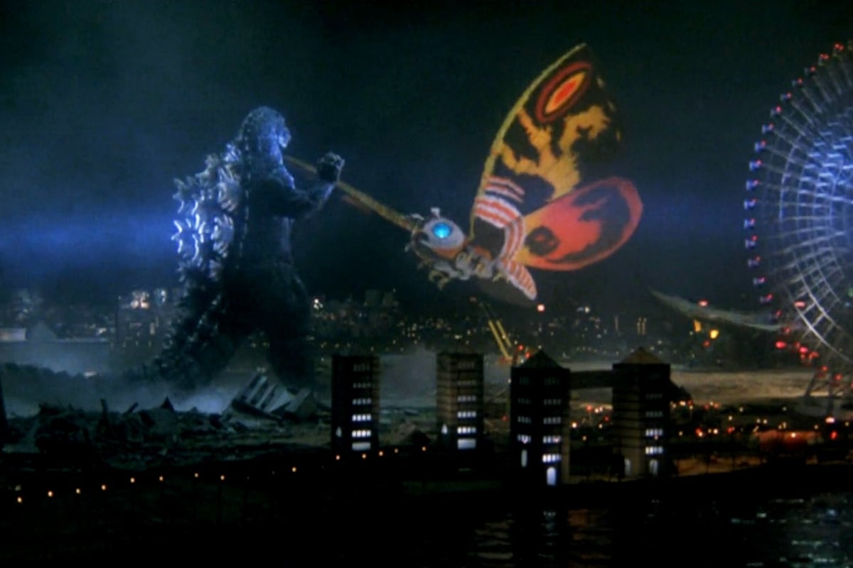 Still from Godzilla and Mothra: Battle for the Earth; Godzilla and Mothra, whose wings are bright orange and yellow, fight against a city lit up in the dark.