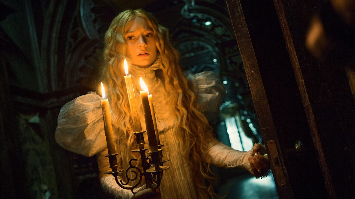 Edith Cushing (Mia Wasikowska) in 'Crimson Peak': a 19th century woman with long blonde hair navigates a dark hallway with a candelabra 