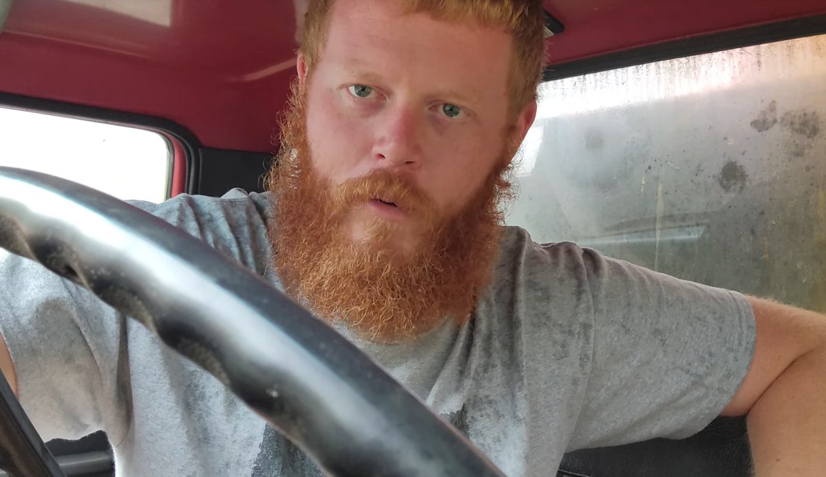 Oliver Anthony, a young white man with a red beard, sits in a truck.