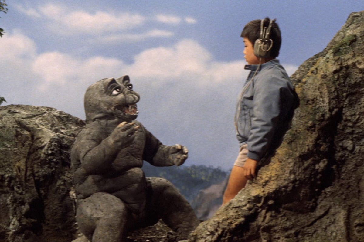 Screenshot from All Monsters Attack; a small, chubby Godzilla type creature faces a small Japanese boy in a rocky island landscape.