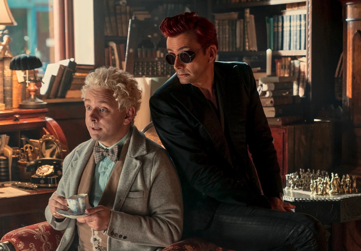 In 'Good Omens' season 2, Aziraphale (Michael Sheen) holds a cup of tea while Crowley (David Tennant) sits behind, wearing sunglasses. They are in Aziraphale's bookshop.