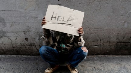 A homeless man sits hopelessly leaning against a wall as there is no one to help him with work and food in his hand holding a sign for help.