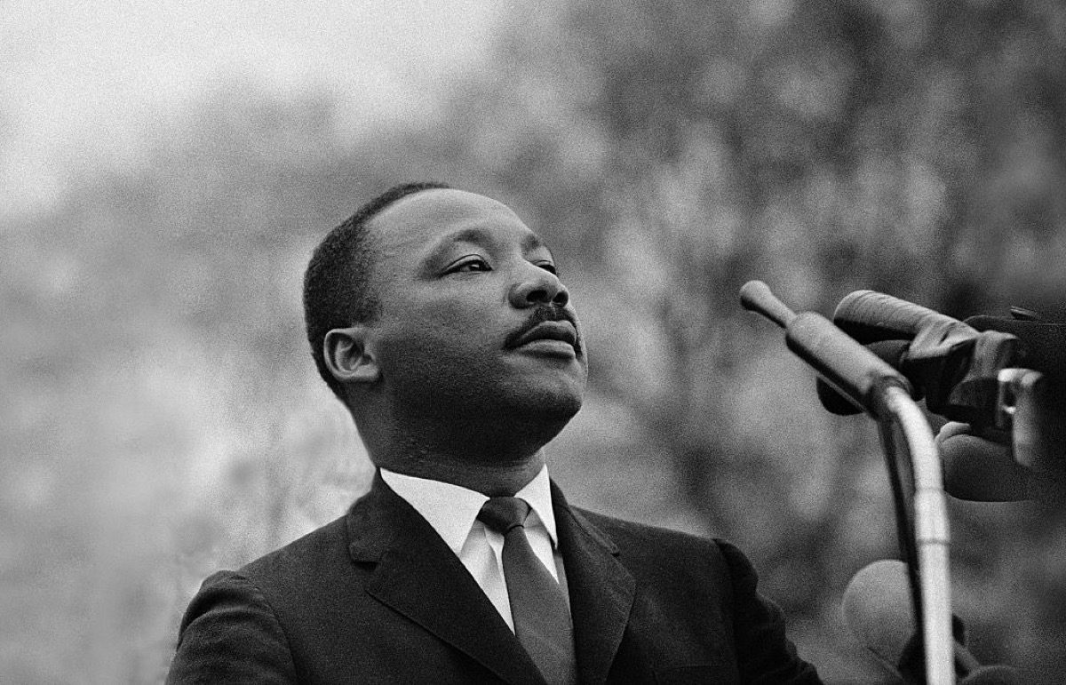 Martin Luther King Jr., standing at a microphone, looking off into the distance with a solemn expression.