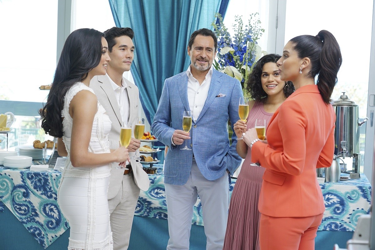 Image of the cast of ABC's 'Grand Hotel' in a scene from the show. It's a group of five people (with Mexican actor Demián Bichir wearing a casual suit with a light blue jacket in the center) standing in a circle in front of a dessert table at a daytime party drinking champagne. Two men and three women. All Latine except for one of the men, who looks like he might be Asian. 