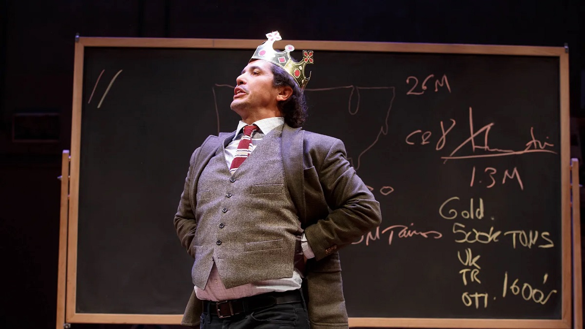 Image of John Leguizamo in a scene from 'Latin History for Morons.' He's a light-skinned Latino standing in front of a blackboard with writing on it wearing a suit jacket, vest, tie, and white buttondown and a gold crown on his head. He has his fists on his hips and stands as if delivering a speech.