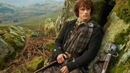 Red haired man wearing a kilt leans against a rock with sprawling landscape behind him in 'Outlander.'