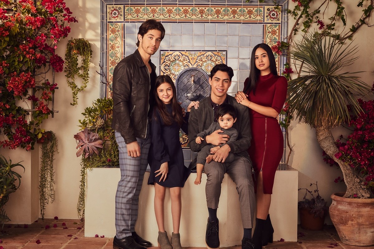 Image of the young cast of Freeform's 'Party of Five' reboot. They are all Latine. Two boys, two girls, and a baby. All of them have dark hair and brown skin. They're standing in front of a tiled fountain surrounded by flowering plants.