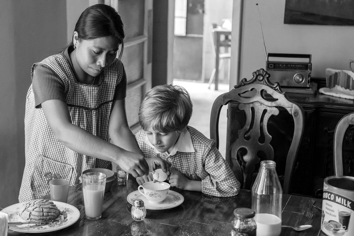 Black and white image of Yalitza Aparicio and a young, white blonde boy in a scene from the film 'Roma.' Aparicio is an Indigenous Mexican woman with long black hair pulled back into a ponytail and wearing a checkered smock over a short sleeved shirt. She is serving the boy breakfast at a table. 