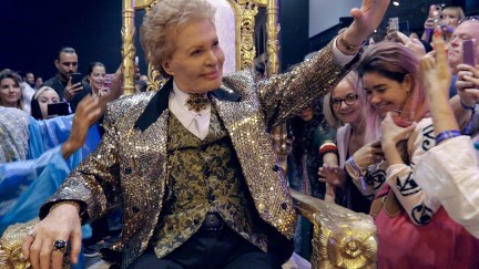 Image of Walter Mercado being brought into a room on a gold throne into a group of cheering fans in a scene from the Netflix documentary 'Mucho, Mucho Amor: The Legend of Walter Mercado.' Walter is a white Puerto Rican man with short wavy blond hair and is wearing a glittery gold blazer over a white button down with a large gold brooch at his throat, a black vest with a gold leaf pattern, and black pants. He's waving to the crowd.