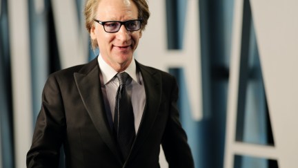 Bill Maher walks, wearing a suit and smiling slightly.